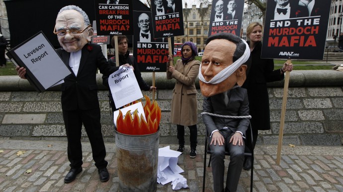 A campaigner wearing a giant mask depicting News Corporation's chairman Rupert Murdoch burns the Leveson report while  another wearing a mask depicting British Prime Minister David Cameron, sits tied to a chair during a protest, calling on MPs to back reform legislation to stop any one media organisation developing a stranglehold over the British media, outside the Queen Elizabeth II Conference Centre in London. (AP Photo/Sang Tan)